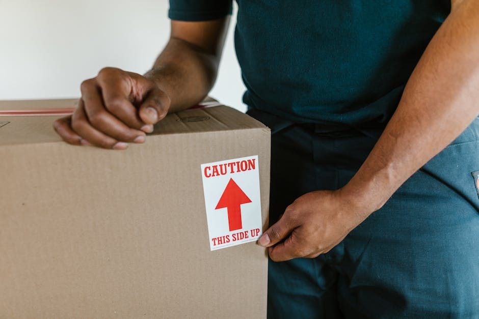 A person wearing dark clothing, including a shirt and trousers, is seen during a home relocation process, holding a large cardboard box with both hands. The box features a red caution label with an upward arrow and the text 'CAUTION THIS SIDE UP,' indicating proper handling during furniture transport and packing. The individual is inside a residential property, standing near a doorway, with a neutral background visible. The scene reflects the loading phase of a furniture transport operation, with the focus on careful handling and the use of packaging materials such as cardboard for packing personal belongings. The process is part of packing and moving services provided by Bromley Man and Van, highlighting the importance of safe loading practices for house removals within the Bromley area.