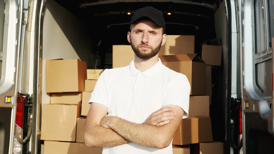 A man with a beard and dressed in a white polo shirt and black cap stands confidently inside a moving van with the rear doors open, surrounded by stacked cardboard boxes of various sizes, some wrapped in plastic. The boxes are arranged on the van's interior floor and walls, indicating a home relocation process. The scene is lit with natural light, highlighting the presence of furniture blankets and lifting equipment such as a trolley nearby. Outside, the van is parked on the pavement, suggesting the loading or unloading phase of a furniture transport and packing operation. This visual exemplifies the steps involved in professional removals services, such as packing, loading, and transporting household belongings, which Bromley Man and Van offers as part of their house removal and logistics services.