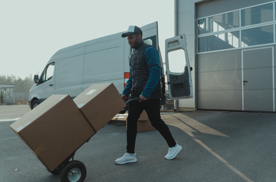 A man with a beard and dressed in a white polo shirt and black cap stands confidently inside a moving van with the rear doors open, surrounded by stacked cardboard boxes of various sizes, some wrapped in plastic. The boxes are arranged on the van's interior floor and walls, indicating a home relocation process. The scene is lit with natural light, highlighting the presence of furniture blankets and lifting equipment such as a trolley nearby. Outside, the van is parked on the pavement, suggesting the loading or unloading phase of a furniture transport and packing operation. This visual exemplifies the steps involved in professional removals services, such as packing, loading, and transporting household belongings, which Bromley Man and Van offers as part of their house removal and logistics services.